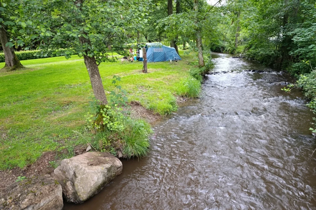 Een blauwe kampeertent op een groen grasveld onder de bomen, direct gelegen aan een kabbelende rivier in de Ardennen.