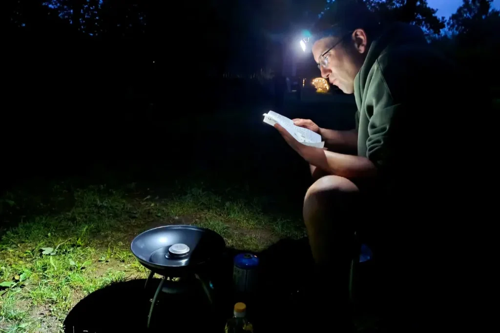 Een man met een hoofdlampje zit in het pikkedonker op een camping op een grasveld en leest aandachtig de instructiehandleiding voor een klein kampeergastoestel dat op de grond voor hem staat.