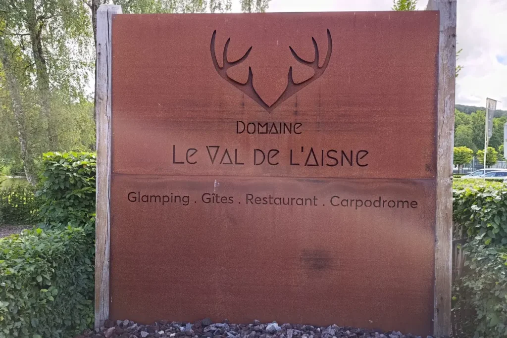 Een groot, roestbruin metalen welkomstbord van Domaine Le Val de l'Aisne met een uitgesneden gewei-logo, omringd door groene hagen.
