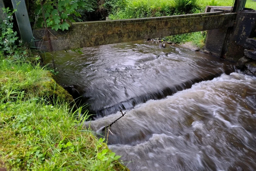 Snelstromend water dat over een kleine stenen verhoging stroomt onder een oude, met mos bedekte houten balk.