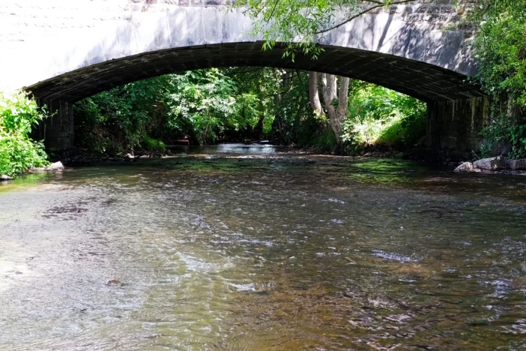 Kijkje vanaf het waterniveau naar een karakteristieke stenen boogbrug over een ondiepe rivier, omringd door groene bomen.