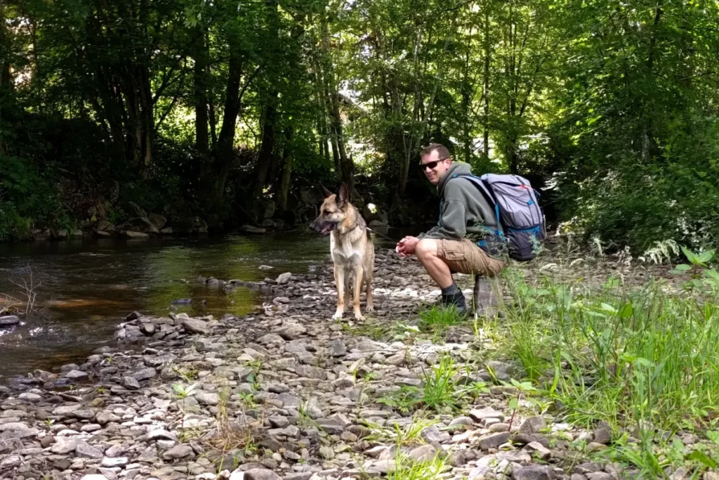 Een man met een rugzak zit gehurkt op een stenen kieuweroever naast zijn Duitse herder Chewey, met op de achtergrond een kabbelende rivier en dicht groen bos.